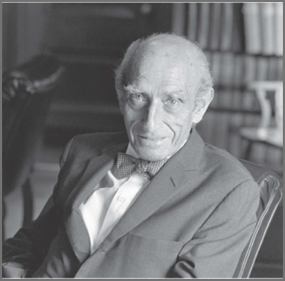 A senior man in a suit and bow tie sitting in an office setting, black and white.